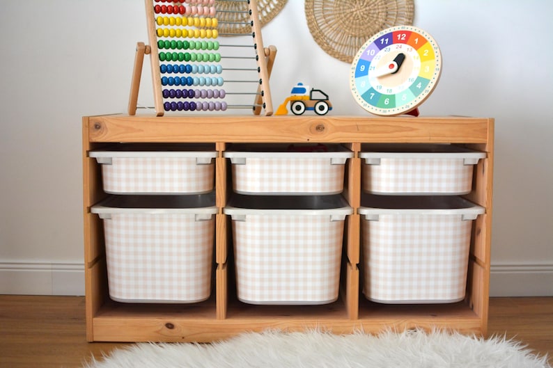 Pu&ograve; includere: Un mobiletto portagiochi in legno chiaro con tre colonne e sei contenitori in plastica bianca con motivo a quadretti beige e bianco. Il mobile ospita un pallottoliere colorato, un piccolo camion giocattolo e un orologio rotondo con numeri da 1 a 24.