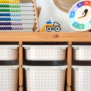 Pu&ograve; includere: Un mobile contenitore in legno chiaro con sei contenitori in plastica bianca con motivo a quadretti beige e bianco. Sopra sono posizionati un abaco in legno con perline colorate, un piccolo veicolo da costruzione giocattolo e un orologio rotondo.