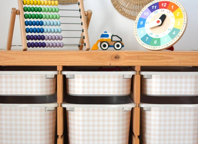 Pu&ograve; includere: Un mobile contenitore in legno chiaro con sei contenitori in plastica bianca con motivo a quadretti beige e bianco. Sopra sono posizionati un abaco in legno con perline colorate, un piccolo veicolo da costruzione giocattolo e un orologio rotondo.