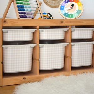 Pu&ograve; includere: Un mobile contenitore in legno chiaro con tre sezioni verticali, ciascuna contenente un contenitore in plastica poco profondo e uno profondo. I contenitori presentano un motivo a quadretti beige e bianco. Il mobile &egrave; rettangolare e aperto.
