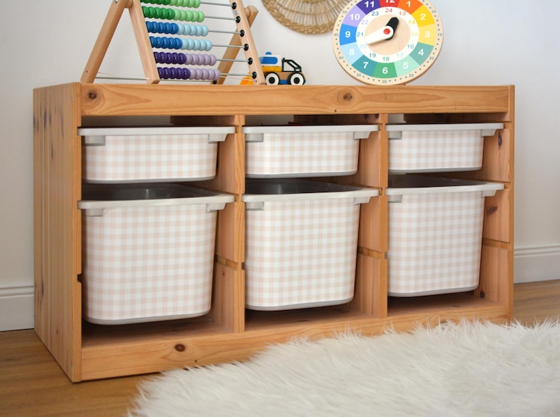 Pu&ograve; includere: Un mobile contenitore in legno chiaro con tre sezioni verticali, ciascuna contenente un contenitore in plastica poco profondo e uno profondo. I contenitori presentano un motivo a quadretti beige e bianco. Il mobile &egrave; rettangolare e aperto.