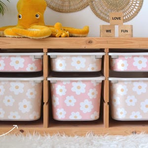 May include: A wooden storage unit with six compartments, each holding a plastic bin. The bins are decorated with a daisy pattern in pink and beige. A yellow octopus plush toy sits on top, along with wooden blocks that spell "WE FUN".