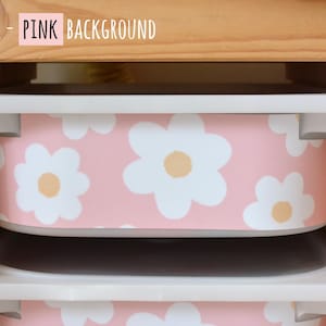 May include: Pink storage bins with a white daisy pattern are stacked in a wooden shelf. The text "SMALL - PINK BACKGROUND" is in the top left corner. The bins have white rims and are designed for organizing items.