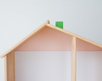 May include: Wooden house-shaped shelf with a light pink interior and a green chimney. The frame is made of natural wood, and the interior is painted a soft pink. The shelf is designed to resemble a house.