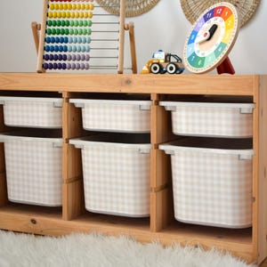 Pu&ograve; includere: Un mobiletto portagiochi in legno chiaro con tre sezioni verticali e sei contenitori in plastica bianca rimovibili. Ogni contenitore &egrave; decorato con un motivo a quadretti beige e bianco. Il mobile &egrave; rettangolare con ripiani aperti.