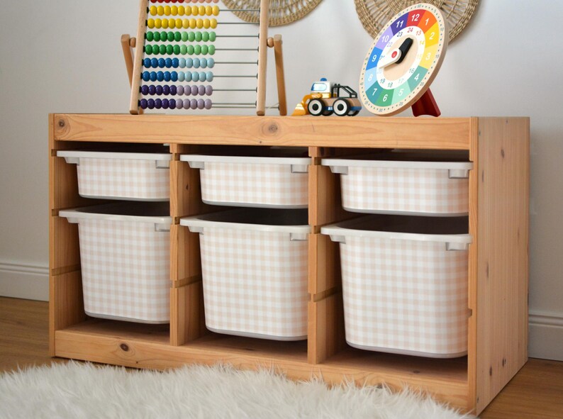 Pu&ograve; includere: Un mobiletto portagiochi in legno chiaro con tre sezioni verticali e sei contenitori in plastica bianca rimovibili. Ogni contenitore &egrave; decorato con un motivo a quadretti beige e bianco. Il mobile &egrave; rettangolare con ripiani aperti.