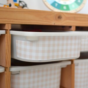 Pu&ograve; includere: Un mobile contenitore in legno chiaro dotato di diversi contenitori rettangolari in plastica. Ogni contenitore &egrave; decorato con un adesivo a motivo vichy beige e bianco sul pannello frontale. L'unit&agrave; presenta un design a scaffali aperti.