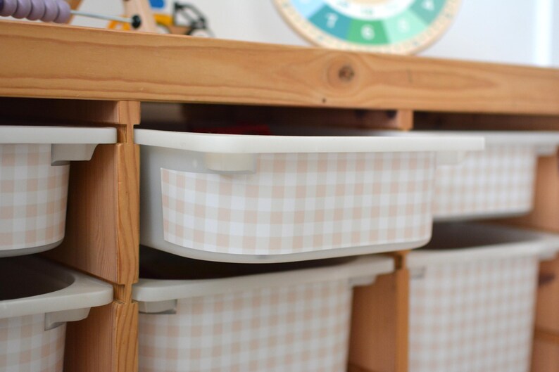 Pu&ograve; includere: Un mobile contenitore in legno chiaro dotato di diversi contenitori rettangolari in plastica. Ogni contenitore &egrave; decorato con un adesivo a motivo vichy beige e bianco sul pannello frontale. L'unit&agrave; presenta un design a scaffali aperti.