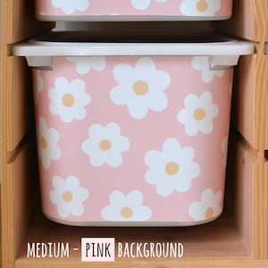 May include: Pink storage bin with a white daisy pattern and a light yellow center. The bin has a white rim and is labeled "MEDIUM - PINK BACKGROUND". The bin is sitting in a wooden shelf.
