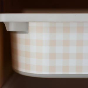 Pu&ograve; includere: Un contenitore rettangolare in plastica con un motivo a quadretti vichy color beige chiaro e bianco. Il contenitore presenta un bordo superiore liscio e piatto con angoli arrotondati, posizionato all'interno di una struttura in legno.
