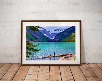 May include: A framed photograph of a mountain lake with turquoise water, surrounded by snow-capped mountains and lush green trees. The lake is calm and reflective, with a few logs floating on the surface.