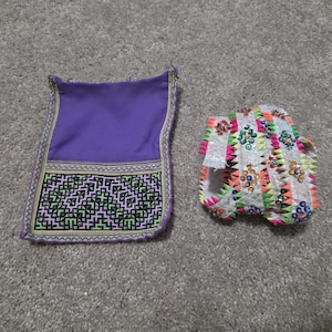 May include: Two pieces of traditional textile art. One is a purple rectangular piece with a geometric patterned border. The other is a white, open-toed shoe-like item with colorful sequin and trim details.