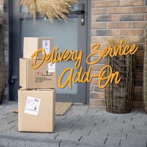 May include: Three stacked cardboard boxes with shipping labels and the text "Delivery Service Add-On" in orange script. The boxes are on a gray stone surface in front of a door and brick wall.