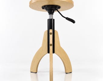 Könnte beinhalten: Ein Hocker mit einer runden Sitzfläche aus hellem Holz und einer schwarzen Metallsäule. Das Untergestell besteht aus drei Beinen aus hellem Holz, die mit goldfarbenen Schrauben an der Säule befestigt sind. Ein schwarzer Hebel sitzt unter dem Sitz.