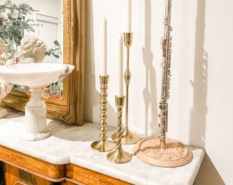 May include: Three gold candle holders with white candles on a white marble surface. A silver flute is on a wooden stand to the right of the candles.