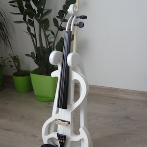 May include: A white electric violin with a black fingerboard and a bow. The violin is displayed on a matching white stand. The instrument is set against a neutral background with green potted plants.