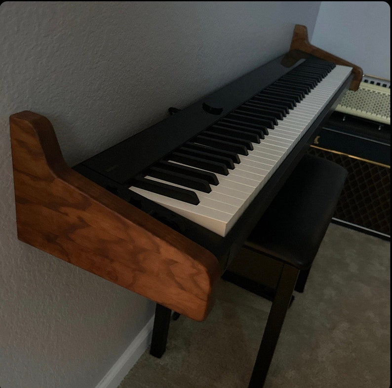 K&ouml;nnte beinhalten: Ein schwarzes digitales Klavier mit wei&szlig;en Tasten, das an einer Wand mit einer Holzhalterung befestigt ist. Das Klavier steht auf einer schwarzen Klavierbank.