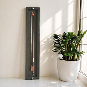 Pode incluir: Um arco de violino exibido em uma moldura retangular cinza escuro. O arco é marrom com uma corda branca e é montado verticalmente. Uma planta em vaso branco fica perto de uma janela ao fundo.