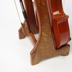 May include: A wooden violin stand with a violin and bow. The stand is made of light brown wood with a natural grain pattern. The violin is a warm brown color with a glossy finish. The bow is white with a dark brown handle.