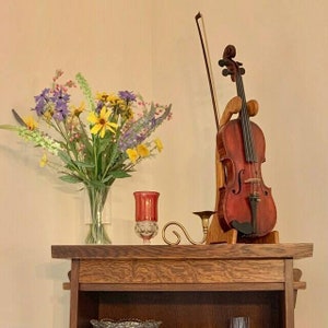 May include: A wooden shelf displays a violin in a wooden stand, with a bow. A bouquet of colorful flowers in a clear vase and a red candle holder are also on the shelf. The violin is a reddish-brown color.