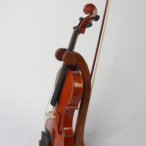 May include: A wooden violin stand with a violin and bow. The stand is made of dark wood and has a curved design. The violin is a warm brown color and is resting in the stand. The bow is leaning against the violin.