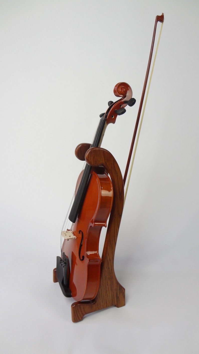 May include: A wooden violin stand with a violin and bow. The stand is made of dark wood and has a curved design. The violin is a warm brown color and is resting in the stand. The bow is leaning against the violin.
