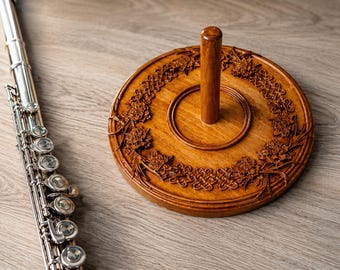 May include: A silver-colored metal flute with multiple keys and a cylindrical body positioned next to a round, medium-brown wooden stand featuring a central vertical post and intricate carved floral and knotwork patterns along the circular rim.
