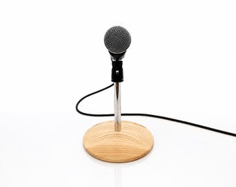 May include: A black dynamic microphone with a silver mesh grille is mounted on a vertical chrome stand. The stand is attached to a circular light-coloured wooden base with a visible grain pattern. A black audio cable extends from the microphone.