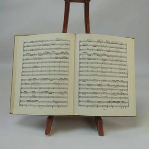 May include: An open book of sheet music displayed on a wooden music stand. The book is brown with cream-colored pages filled with musical notation. The stand is a dark brown color and has a simple design.