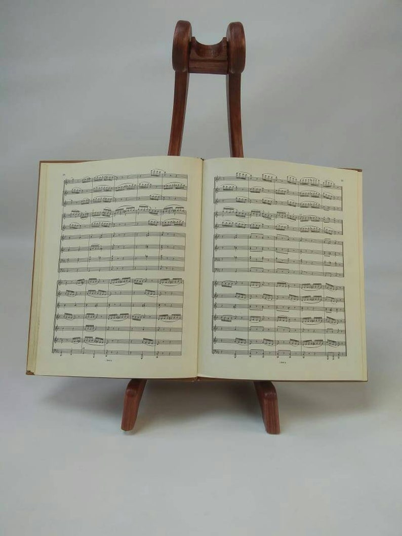 May include: An open book of sheet music displayed on a wooden music stand. The book is brown with cream-colored pages filled with musical notation. The stand is a dark brown color and has a simple design.