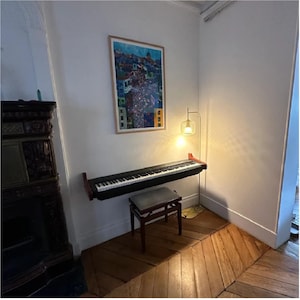 K&ouml;nnte beinhalten: Ein schwarzes digitales Klavier mit einem Holzst&auml;nder ist an einer Wand in einem Raum mit wei&szlig;en W&auml;nden und Holzb&ouml;den montiert. Eine Stehlampe mit einem Glasschirm steht in der Ecke des Raumes. Ein gerahmter Druck einer bunten Stadtlandschaft h&auml;ngt an der Wand.