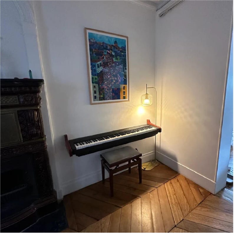 K&ouml;nnte beinhalten: Ein schwarzes digitales Klavier mit einem Holzst&auml;nder ist an einer Wand in einem Raum mit wei&szlig;en W&auml;nden und Holzb&ouml;den montiert. Eine Stehlampe mit einem Glasschirm steht in der Ecke des Raumes. Ein gerahmter Druck einer bunten Stadtlandschaft h&auml;ngt an der Wand.