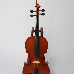 May include: A varnished wooden violin with a bow, displayed on a wooden stand. The violin has a warm, reddish-brown finish with black strings and a black chin rest. The bow is positioned vertically.