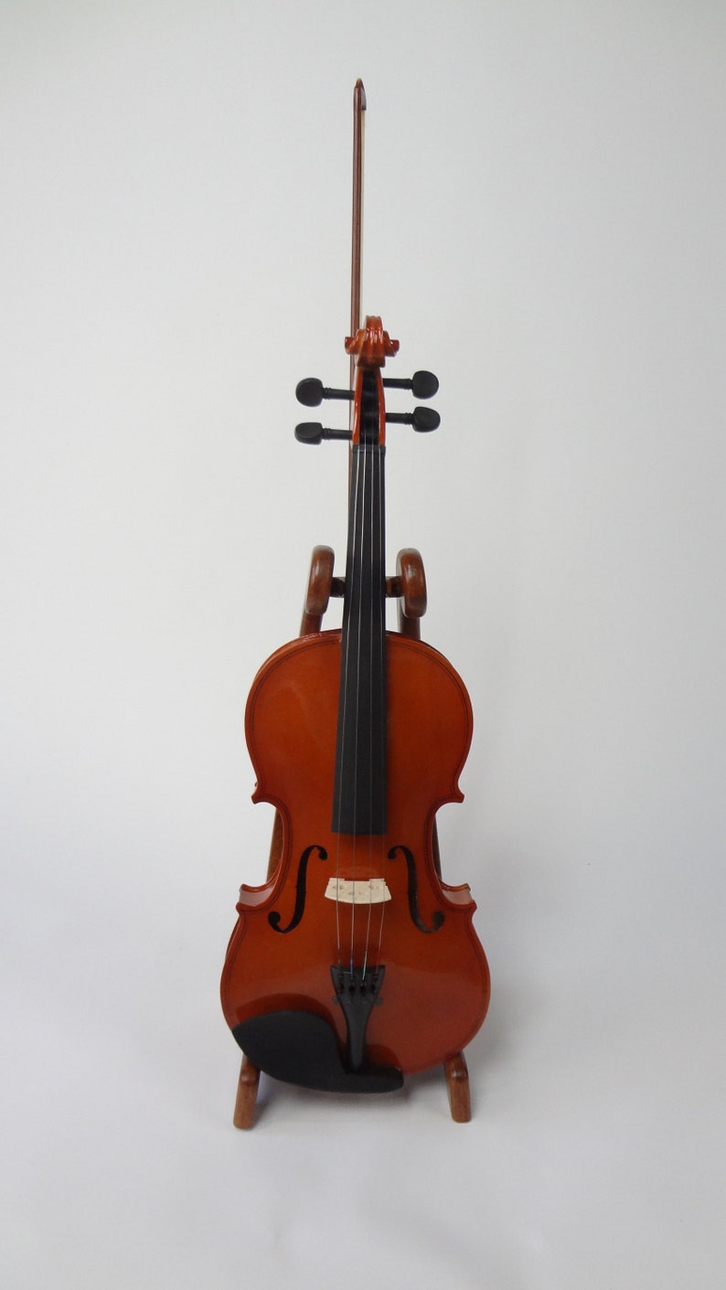 May include: A varnished wooden violin with a bow, displayed on a wooden stand. The violin has a warm, reddish-brown finish with black strings and a black chin rest. The bow is positioned vertically.