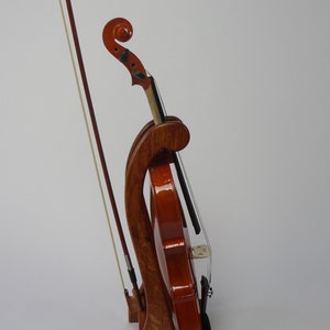 May include: A wooden violin stand with a violin and bow. The stand is made of dark wood and has a curved design. The violin is a warm brown color and is held in place by the stand. The bow is resting next to the violin.