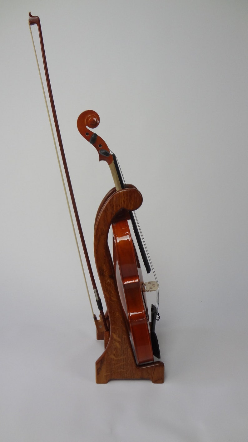 May include: A wooden violin stand with a violin and bow. The stand is made of dark wood and has a curved design. The violin is a warm brown color and is held in place by the stand. The bow is resting next to the violin.