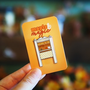 May include: A white enamel pin with a gold outline depicting a small wooden stand with a sign that reads "Welcome to the Pumpkin Patch". The pin is on a bright orange background with the words "maple & magic" in white.