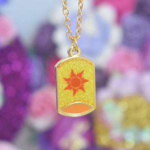 May include: A gold-coloured necklace with a pendant shaped like a yellow lantern. The lantern has a glittery texture and features an orange sun design. The pendant is rectangular with rounded edges.