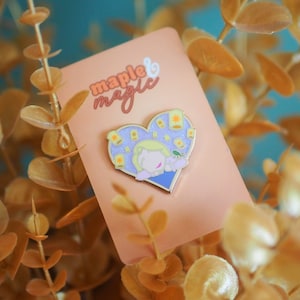 May include: A gold heart-shaped enamel pin with a cartoon image of a girl with blonde hair and a purple dress. The girl is surrounded by small yellow lanterns and the text "maple & magic" is printed on the background.