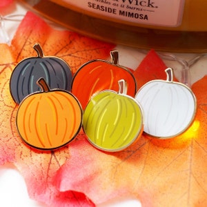 May include: Four enamel pumpkin pins in different colours: orange, green, white, and grey. The pins are on a red and orange autumn leaf background.