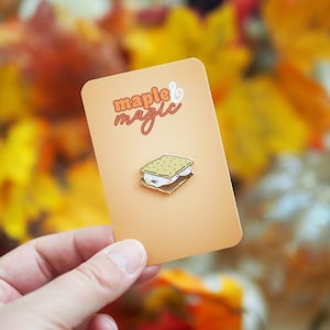 May include: A gold enamel pin featuring a cartoon illustration of a s'more. The pin is on a white card with the text "maple & magic" in orange and brown lettering.