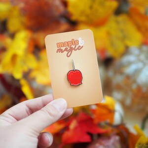 May include: A gold enamel pin featuring a red candy apple with a brown stick. The pin is on a card with the text "maple & magic" in a brown and orange font.