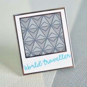 May include: A silver and white enamel pin shaped like a Polaroid photo with a gray and white geometric pattern inside and the text "World traveller" in blue.