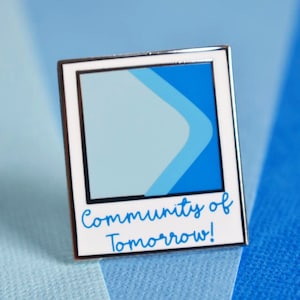 May include: A silver enamel pin with a blue and light blue design inside a white frame. The text "Community of Tomorrow!" is written below the frame.