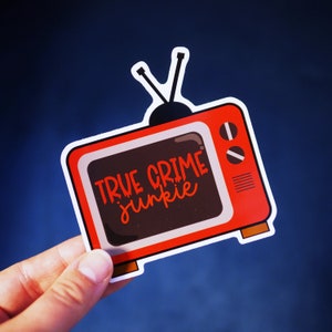 Op de afbeelding: Rode en zwarte sticker in de vorm van een tv met de tekst "True Crime Junkie" in witte letters.