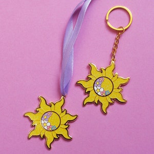 May include: Two gold-tone sun charms with a yellow and glitter finish. One charm is attached to a purple ribbon and the other is attached to a gold-tone key ring. Both charms feature a stylised sun with a half-moon shape and floral details.