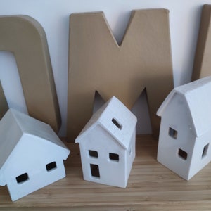 May include: Three white ceramic houses with cut-out windows and doors. The houses are sitting on a wooden surface in front of large cardboard letters that spell "HOME".