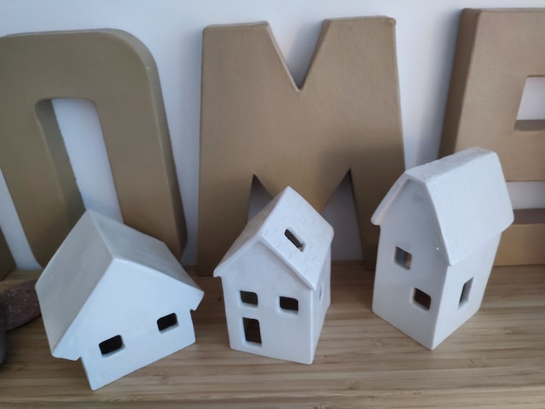 May include: Three white ceramic houses with cut-out windows and doors. The houses are sitting on a wooden surface in front of large cardboard letters that spell "HOME".
