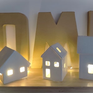 May include: Three white ceramic houses with lit windows, arranged on a wooden shelf in front of gold cardboard letters spelling "HOME".