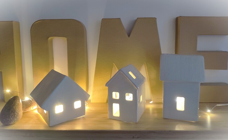 May include: Three white ceramic houses with lit windows, arranged on a wooden shelf in front of gold cardboard letters spelling "HOME".
